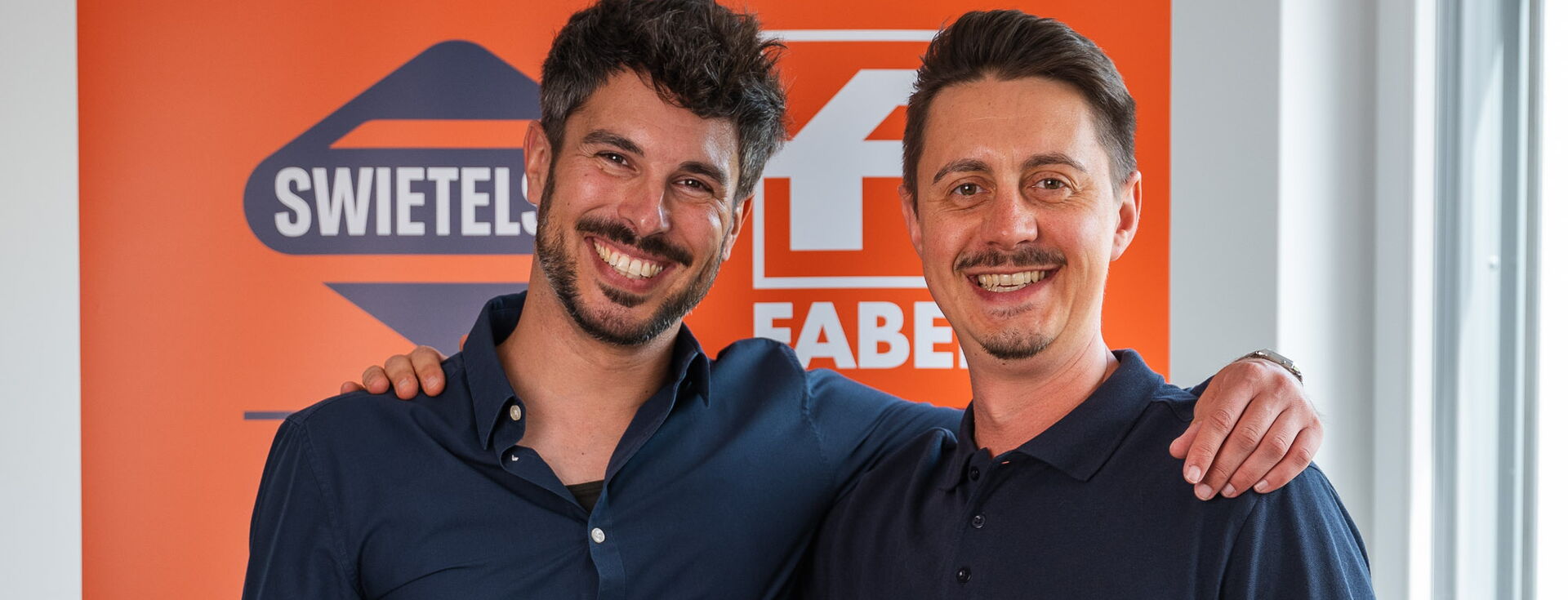 Zwei lächelnde Männer stehen Arm in Arm in einem Raum vor einem orangefarbenen Swiftel-Banner.