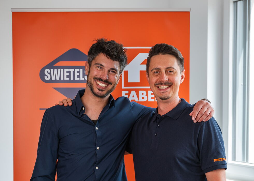 Zwei lächelnde Männer stehen Arm in Arm in einem Raum vor einem orangefarbenen Swiftel-Banner.