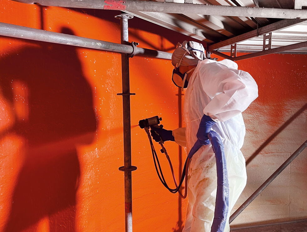 Arbeiter in Schutzanzug sprüht eine leuchtend orange Wand in einer Industrieanlage.