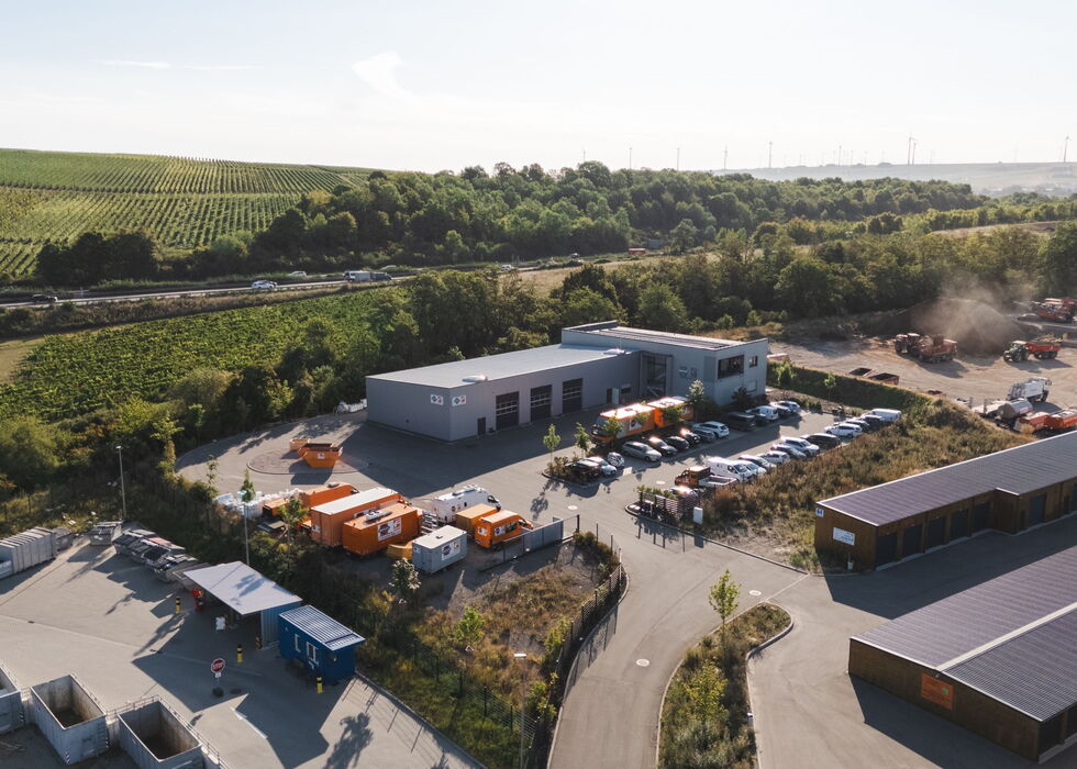 Luftbild eines Industriekomplexes mit Lagerhallen und Fahrzeugen, umgeben von grünen Weinbergen und Feldern.