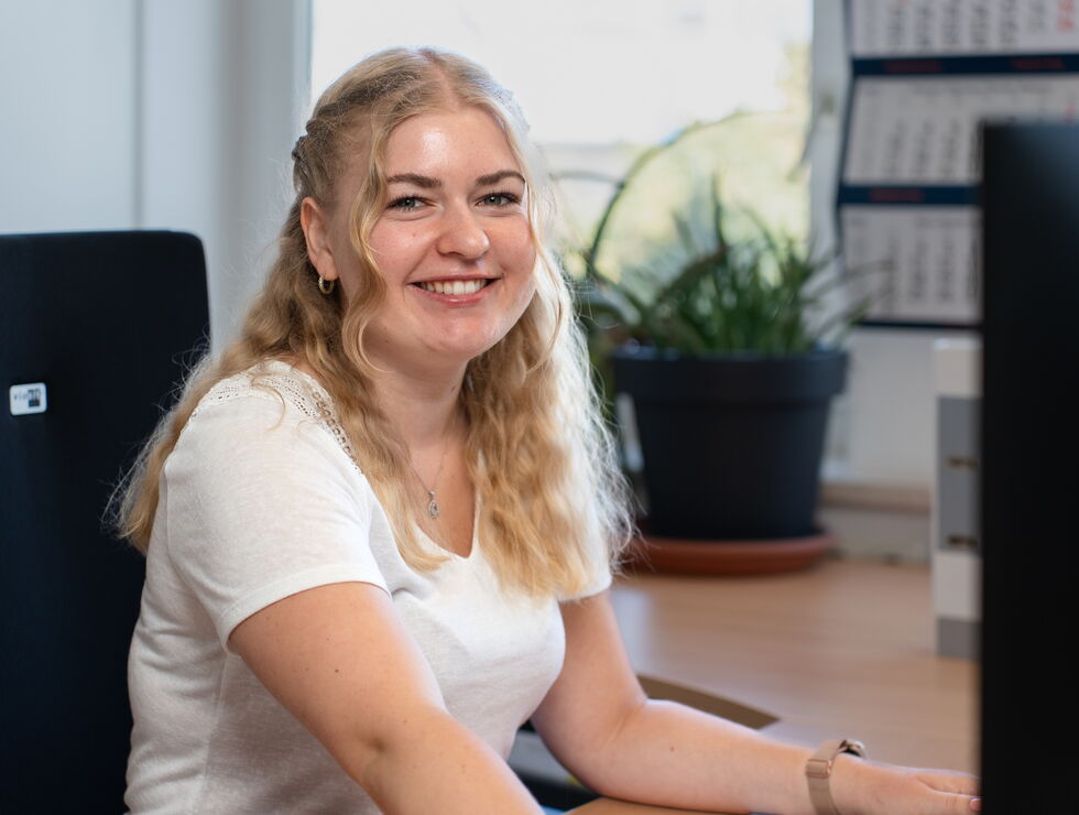 Junge Frau mit blonden Haaren sitzt auf einem Bürostuhl und lächelt, während sie am Computer arbeitet
