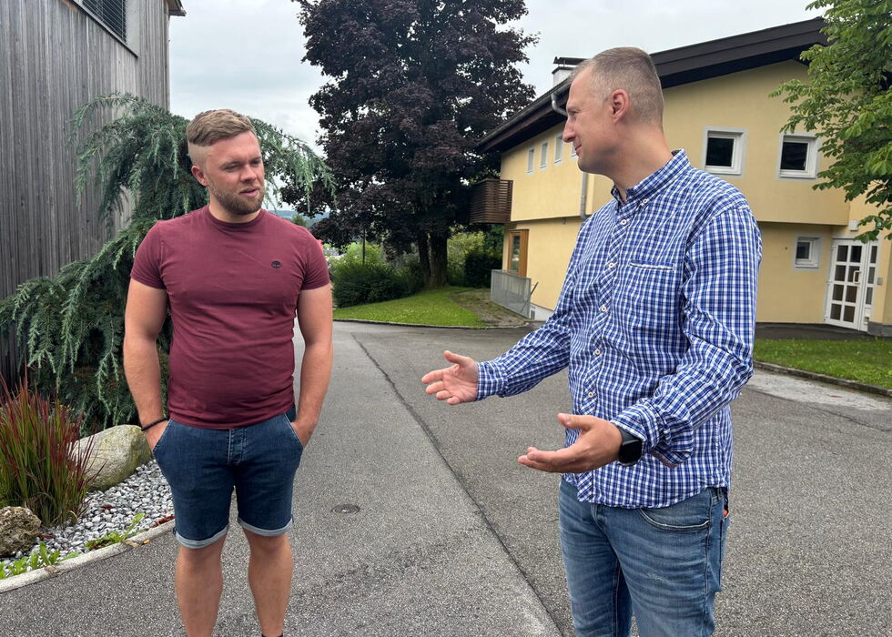 Zwei Männer stehen draußen auf einem Weg und unterhalten sich zwischen Gebäuden und Bäumen.