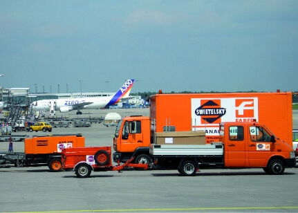 Eine belebte Flughafenszene mit orangefarbenen Servi fahrzeugen und einem Passagierflugzeug im Hintergrund.