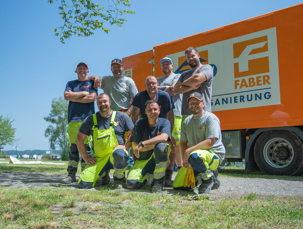 Sieben Bauarbeiter posieren und lächeln vor einem orangen FABER-LKW an einem sonnigen Tag