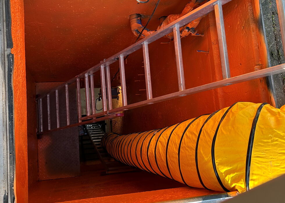Ein Arbeiter klettert eine Leiter in einen leuchtend orangefarbenen Versorgungsschacht hinunter, mit einem großen Belüftungsschlauch.