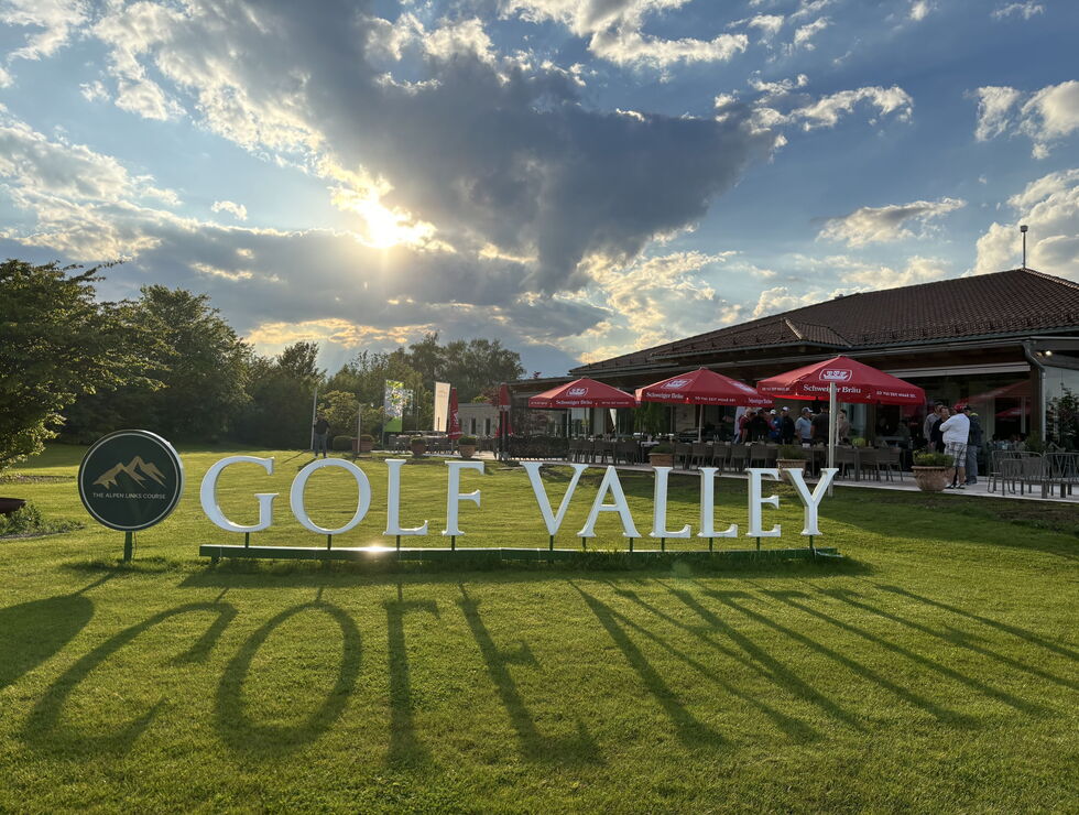 Großes Golf-Valley-Schild auf einer grünen Wiese vor Clubhaus, roten Sonnenschirmen und dramatischem Abendhimmel.