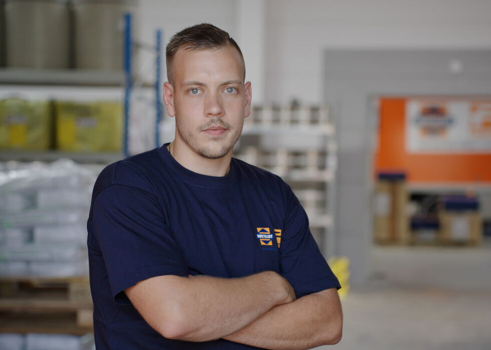Ein Mann steht in einem Lager mit verschränkten Armen vor gestapelten Waren.