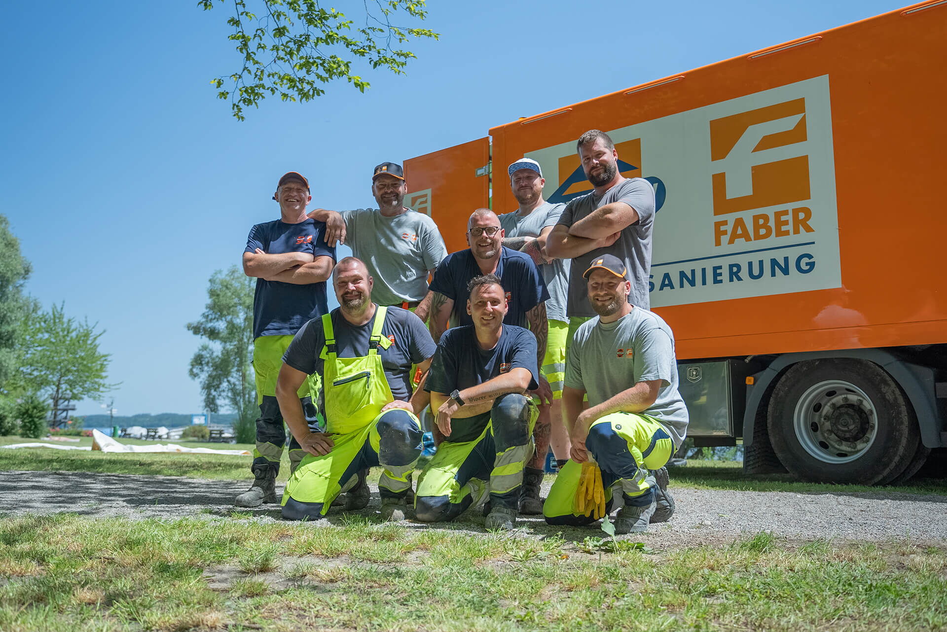 Gruppe von Mitarbeitenden posiert vor einem orangefarbenen Lkw im Freien bei Sonnenschein.