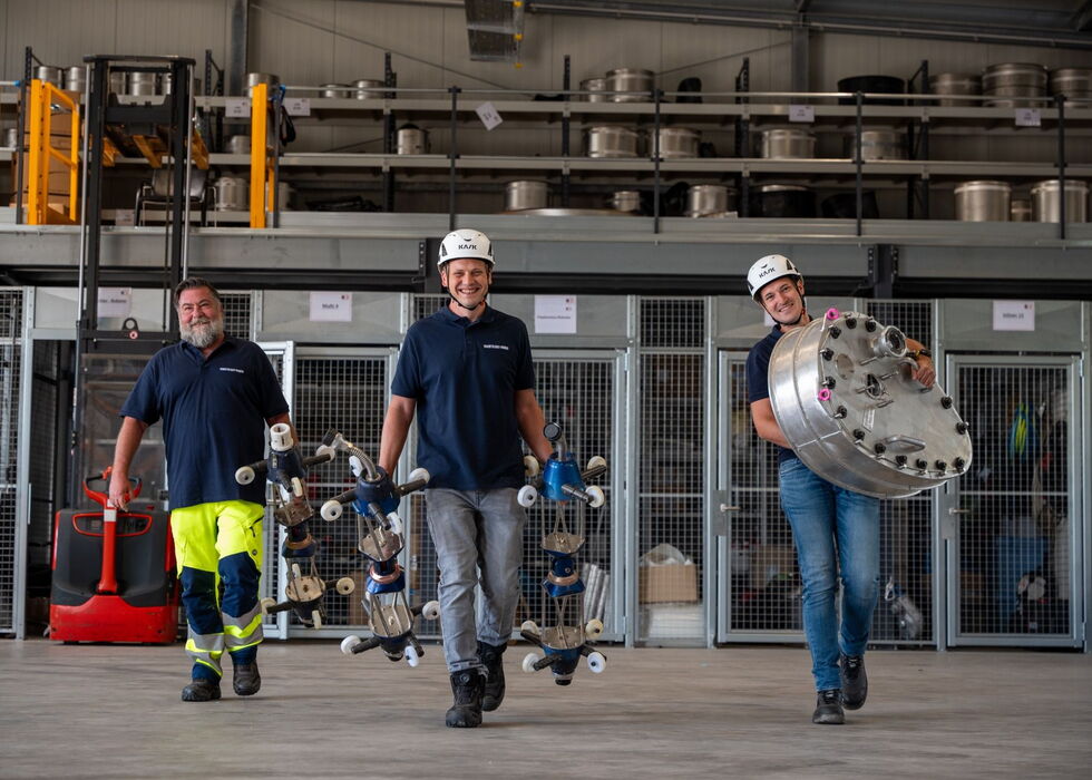 Drei Techniker in Arbeitskleidung und Helmen stehen in einer Werkstatt und halten mechanische Komponenten sowie ein großes rundes Teil.