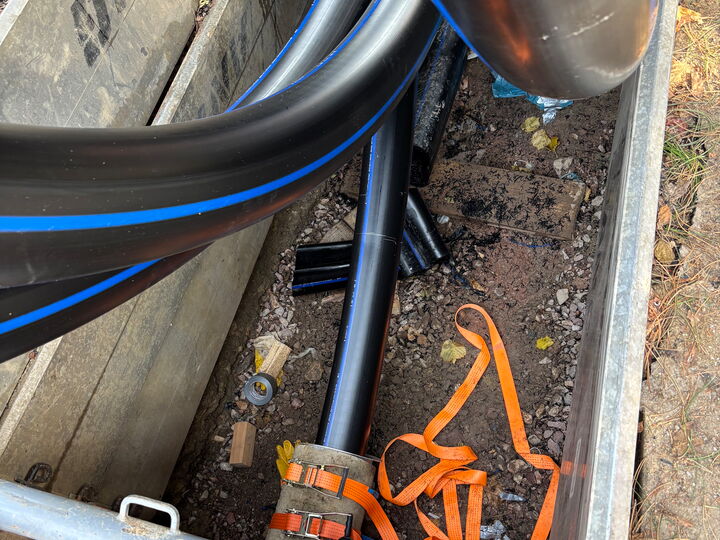 Schwarze Versorgungsleitungen liegen in einem Graben, mit orangefarbenen Gurten und Bauschutt.