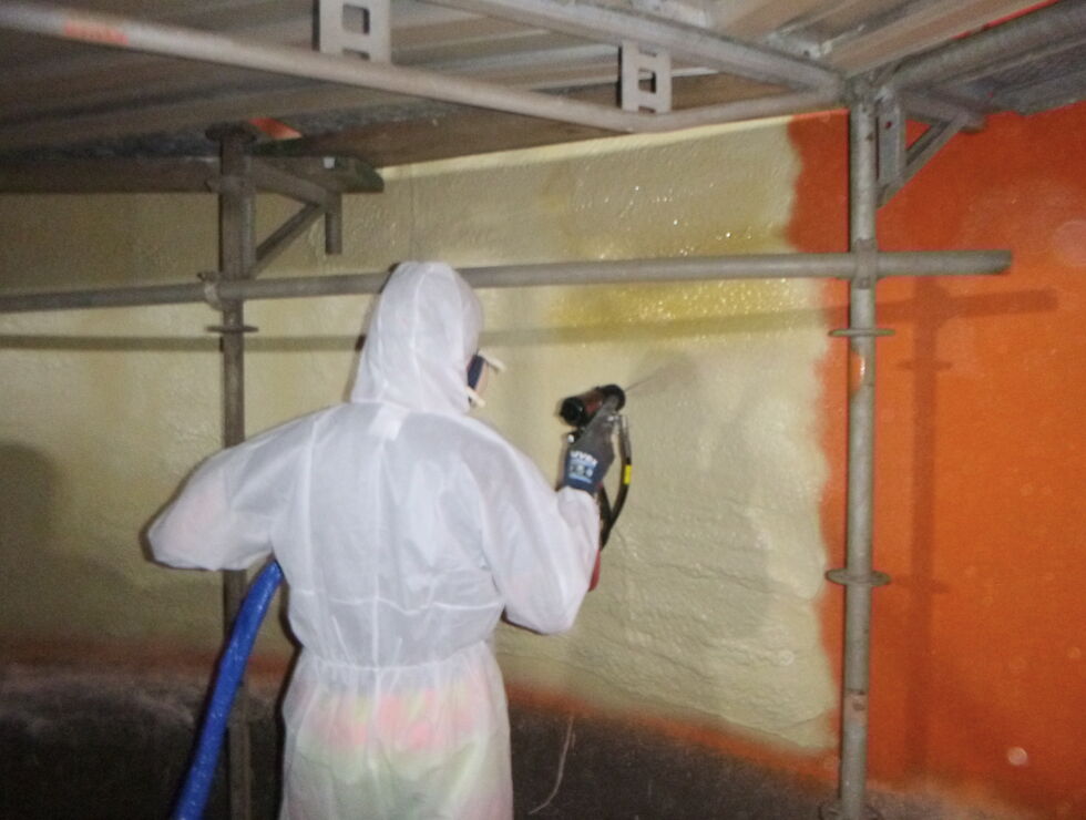 Arbeiter in Schutzanzug sprüht Beschichtung auf eine Wand im Stahlbehälter.