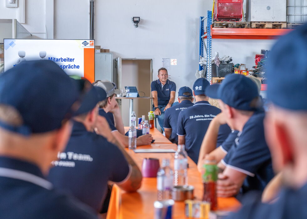 Arbeiter sitzen an langen Tischen und hören in einer Industriehalle einer Besprechung zu.