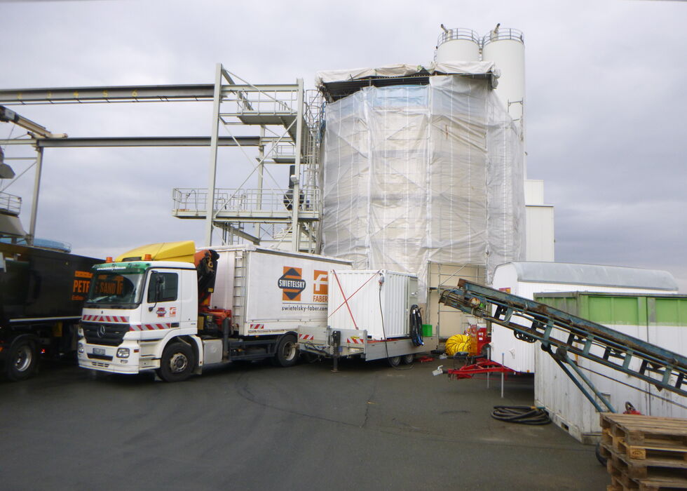 Lkw und Förderanlage neben einer eingewickelten Silostruktur auf einem Industriegelände.
