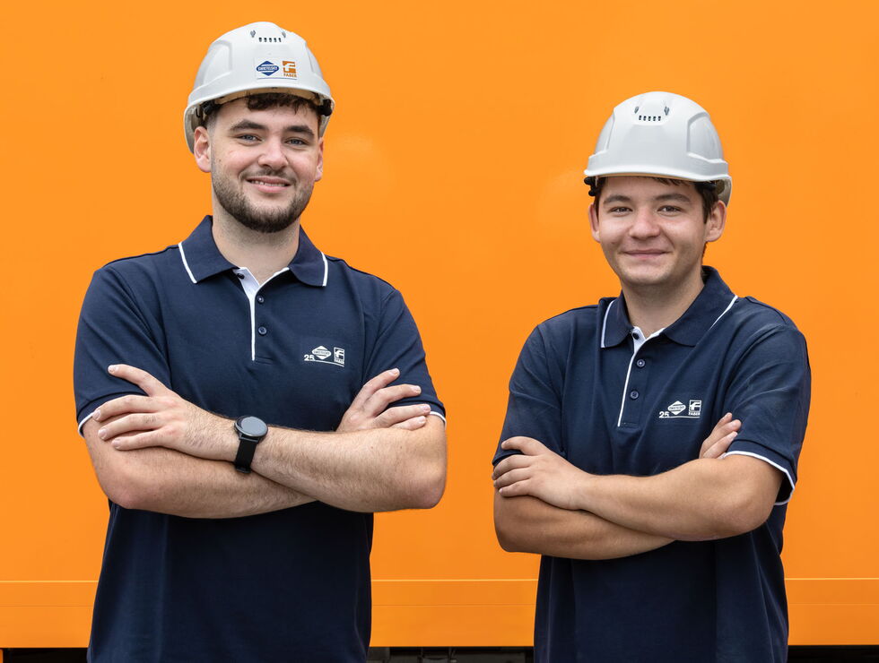 Zwei lächelnde Bauarbeiter mit Schutzhelmen und dunkelblauen Poloshirts stehen mit verschränkten Armen vor orangefarbenem Hintergrund.