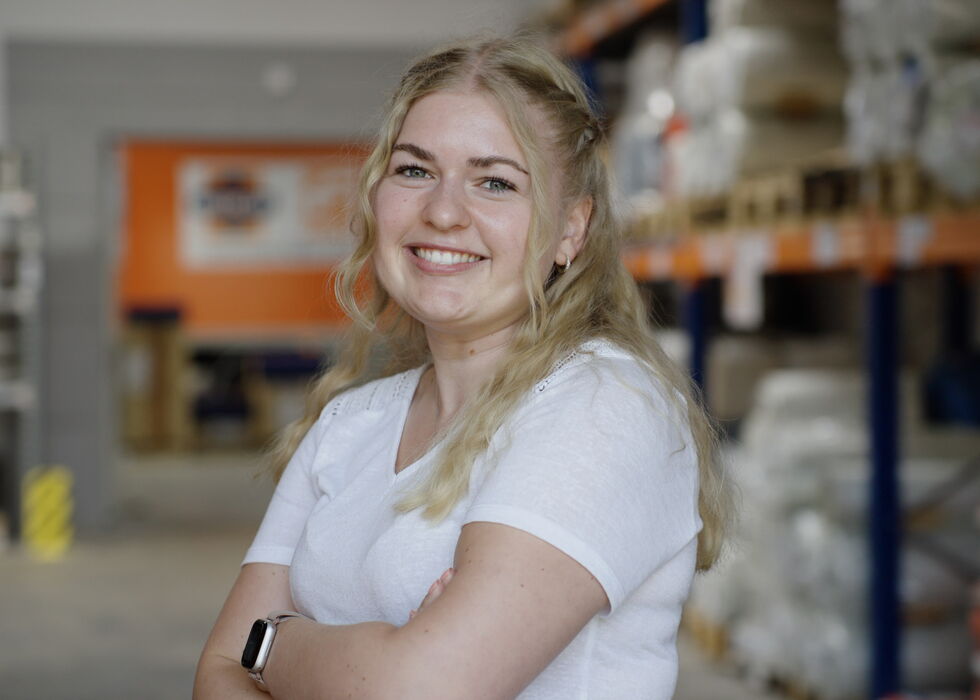 Lächelnde blonde Frau in weißem Shirt, stehend in einem Laden mit Regalen im Hintergrund.