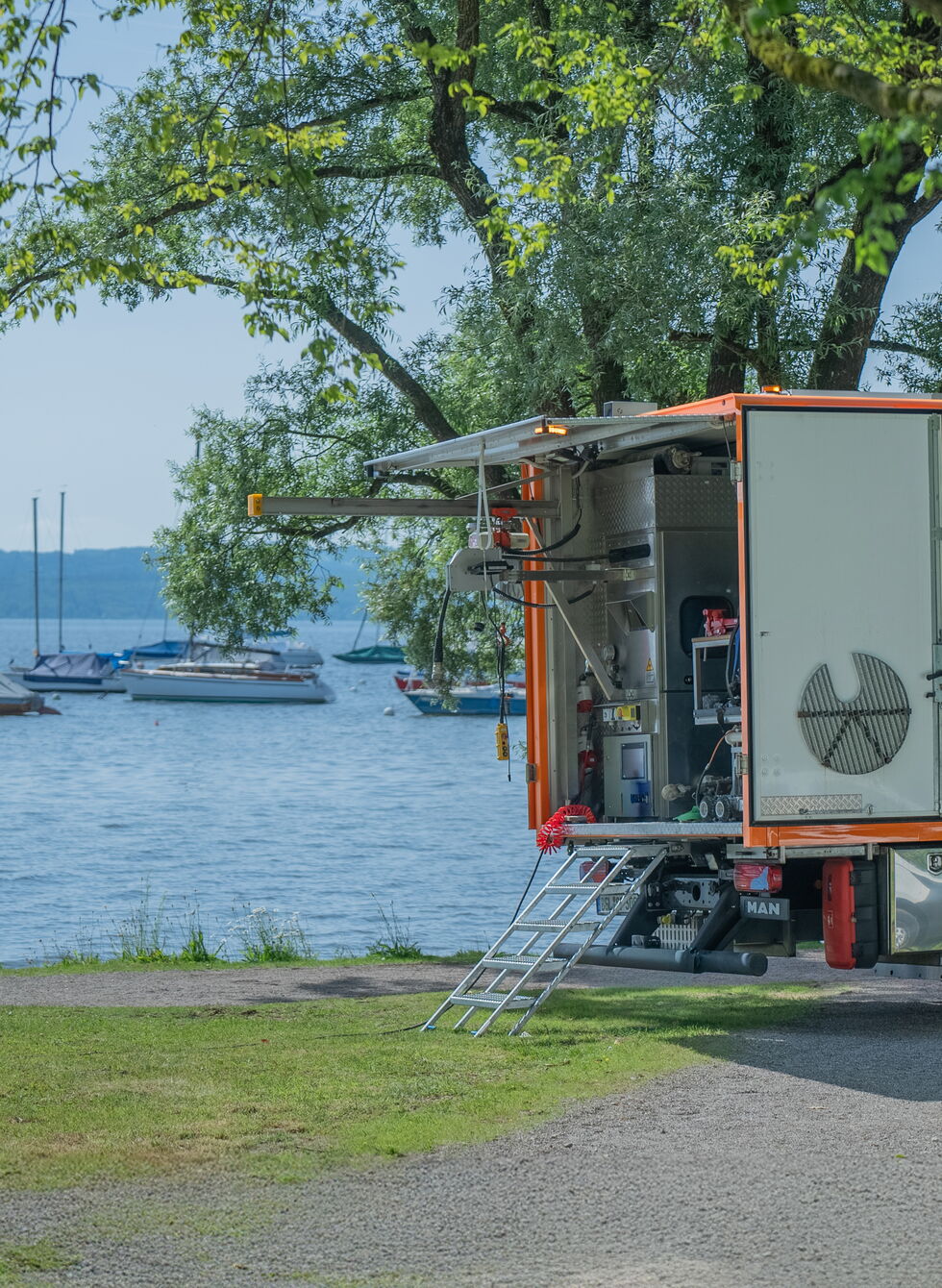 Orange ice cream truck parked beside a lake with sailboats in the background and trees overhead.