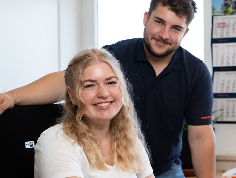 Lächende junge Frau sitzt am Schreibtisch und benutzt Computermaus, ein Mann steht hinter ihr in einer lockeren Büroumgebung