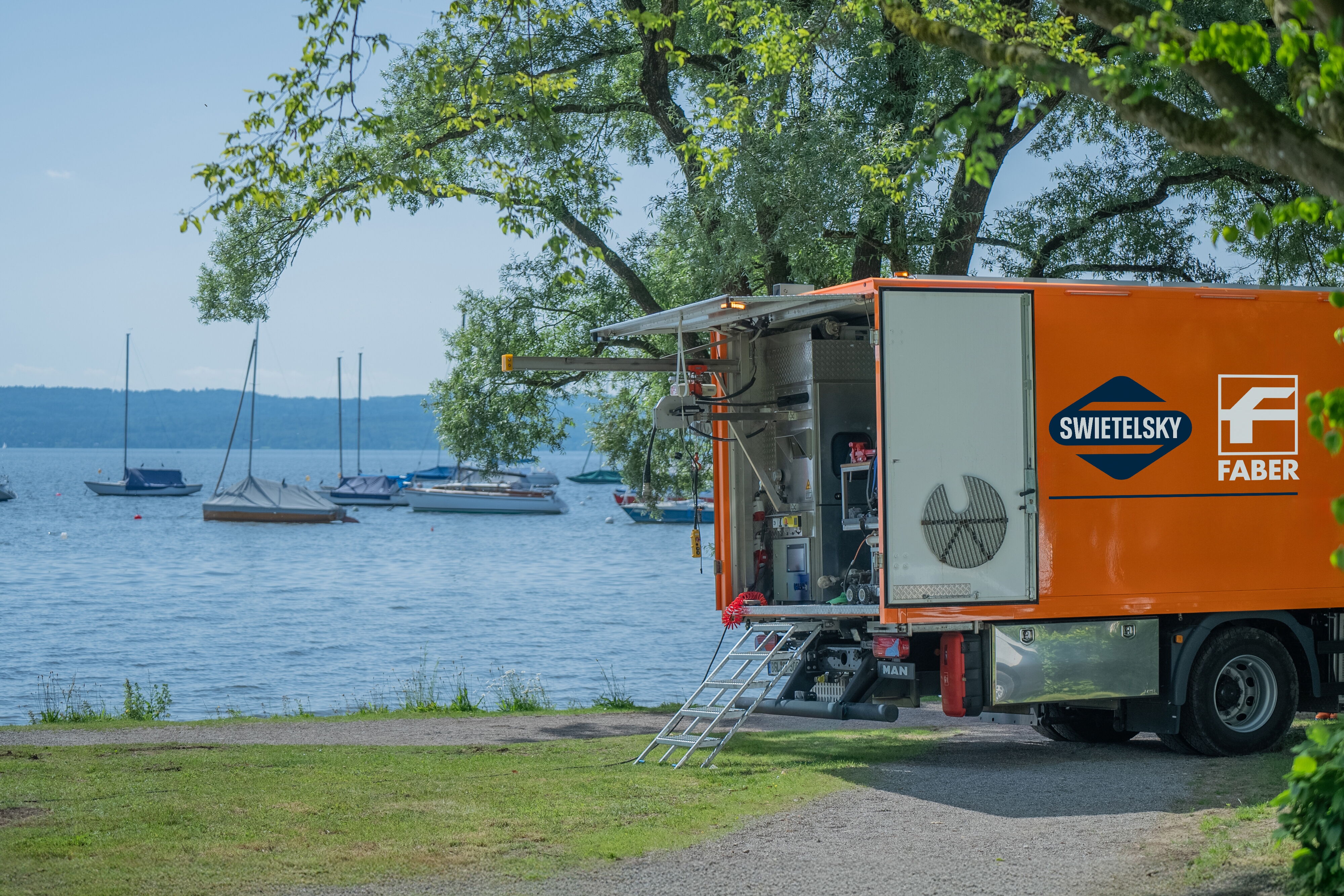 Oranger Faber-Lkw am Seeufer mit Segelbooten und Bäumen im Hintergrund.