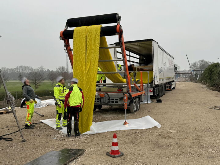 Arbeiter in Warnkleidung entladen eine große gelbe Geberolle neben einer Landstraße aus einem Lkw.