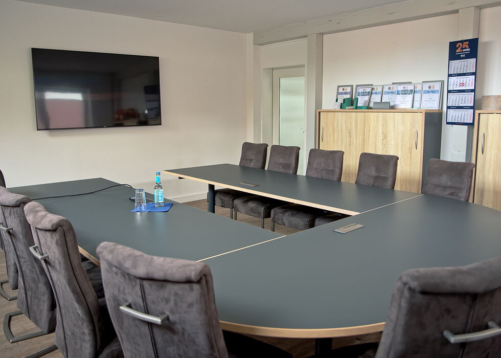 A conference room with a large U-shaped meeting table, upholstered chairs, a wall-mounted TV, and cabinets in the background.