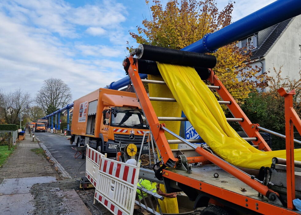 Orangefarbene Hubarbeitsbühne mit gelber Plane und Sprühdüse auf einer Straße neben Gebäuden.