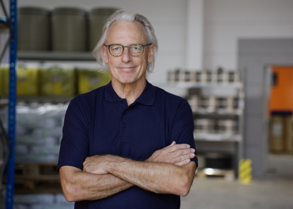 Lächelnder älterer Mann mit Brille steht mit verschränkten Armen in einer Lager- oder Industrieumgebung.