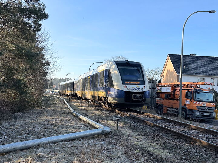 Ein moderner Personenzug fährt auf Bahngleisen vorbei an Häusern und Wartungsfahrzeugen, links daneben ist eine Entwässerungsleitung zu sehen.