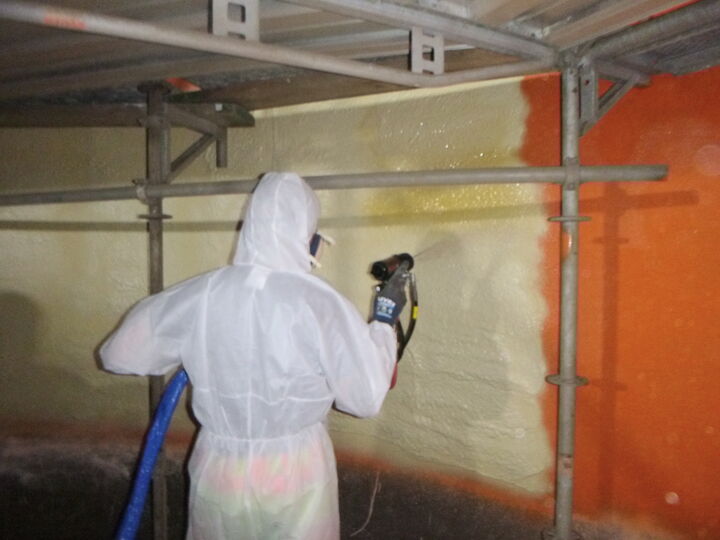 Arbeiter in Schutzanzug sprüht Beschichtung auf eine Wand im Stahlbehälter.