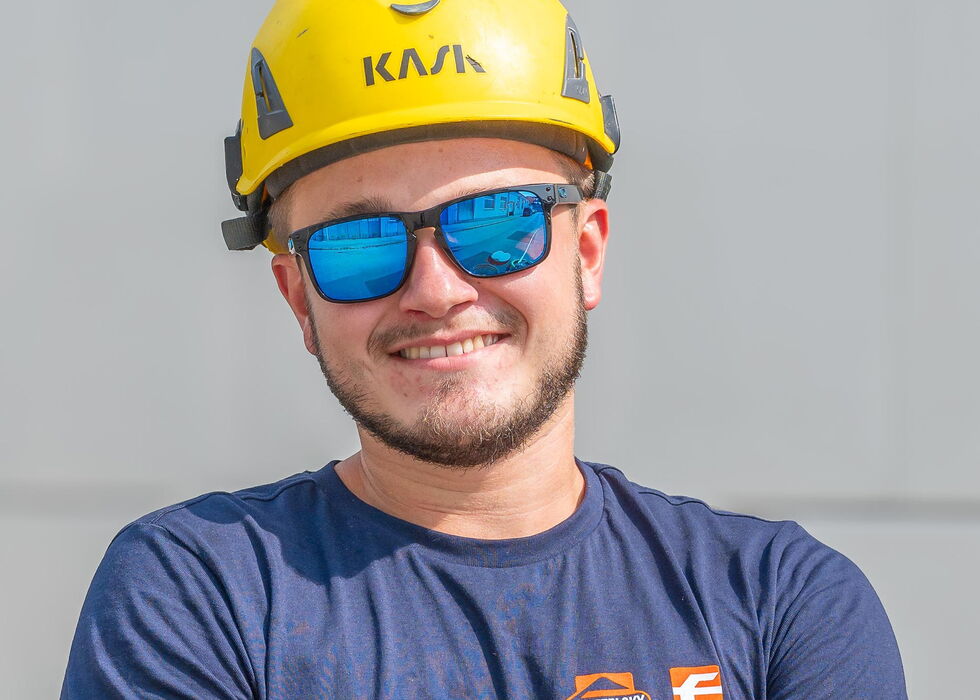 Lächelnder Bauarbeiter mit gelbem Helm und blauer Sonnenbrille, die Arme verschränkt.