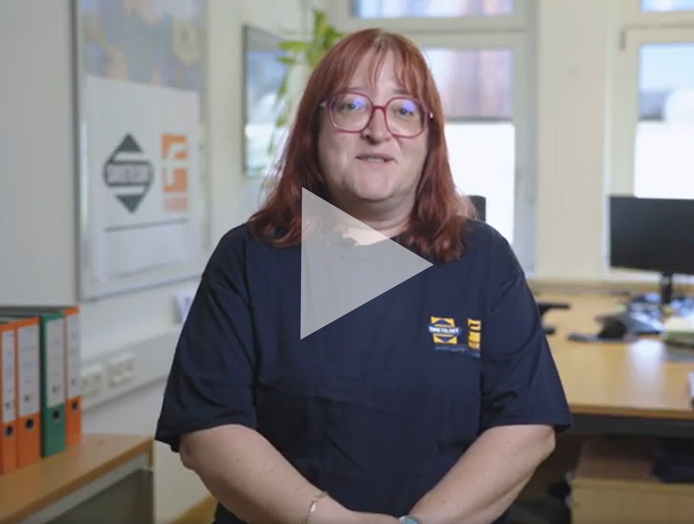 Eine Frau mit roten Haaren und Brille trägt ein dunkles T-Shirt und steht in einem hellen Büro mit Tischen und Computern im Hintergrund.