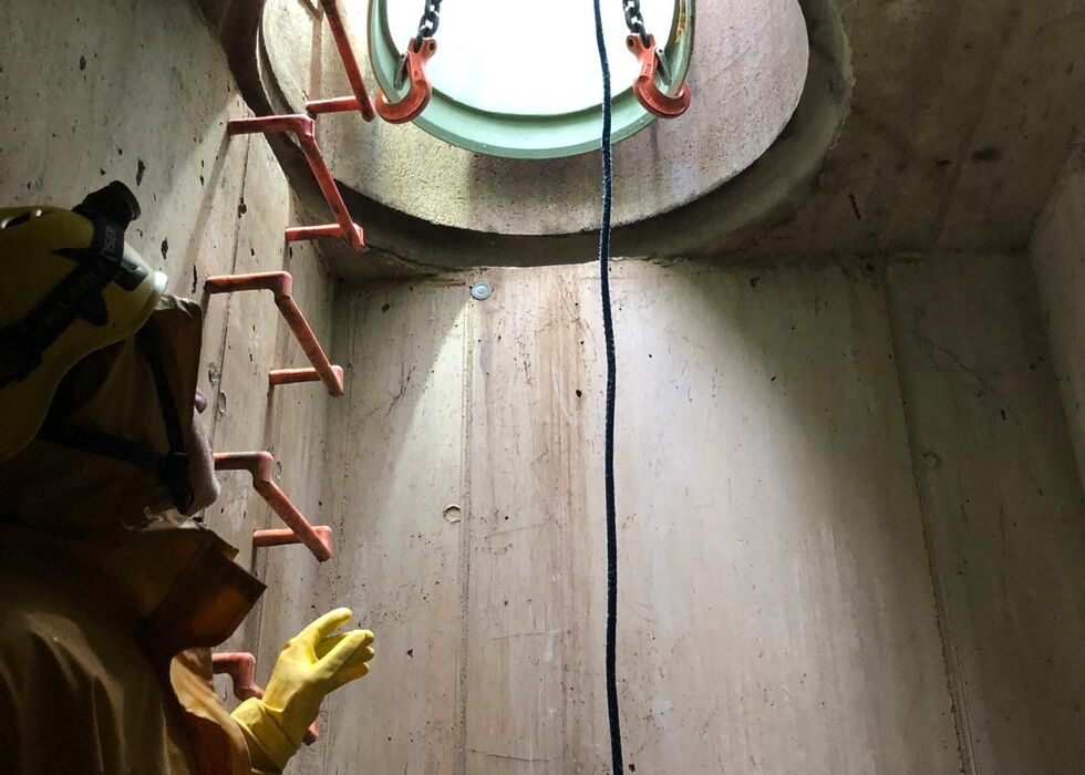 Ein Arbeiter in Schutzkleidung klettert eine Leiter in einem Betonschacht hinauf zu einer runden Öffnung mit Hebeketten.