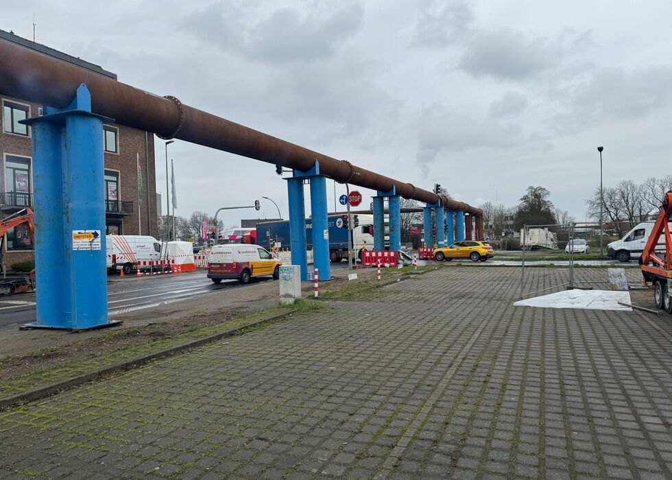 Große rostige Pipeline auf blauen Stützen neben einem gepflasterten Bereich und Straßenarbeiten an einem bewölkten Tag.