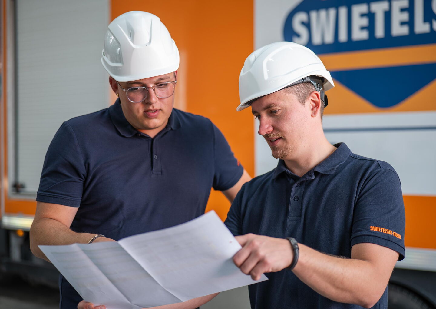Zwei Männer mit weißen Helmen betrachten Baupläne vor einem Gebäude mit Swietelsky-Logo