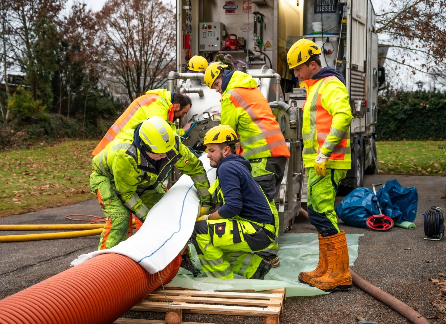Eine Gruppe von Arbeitern in gelber Schutzkleidung, die an einer Wartungsaufgabe mit großen Geräten und Schläuchen zusammenarbeitet.