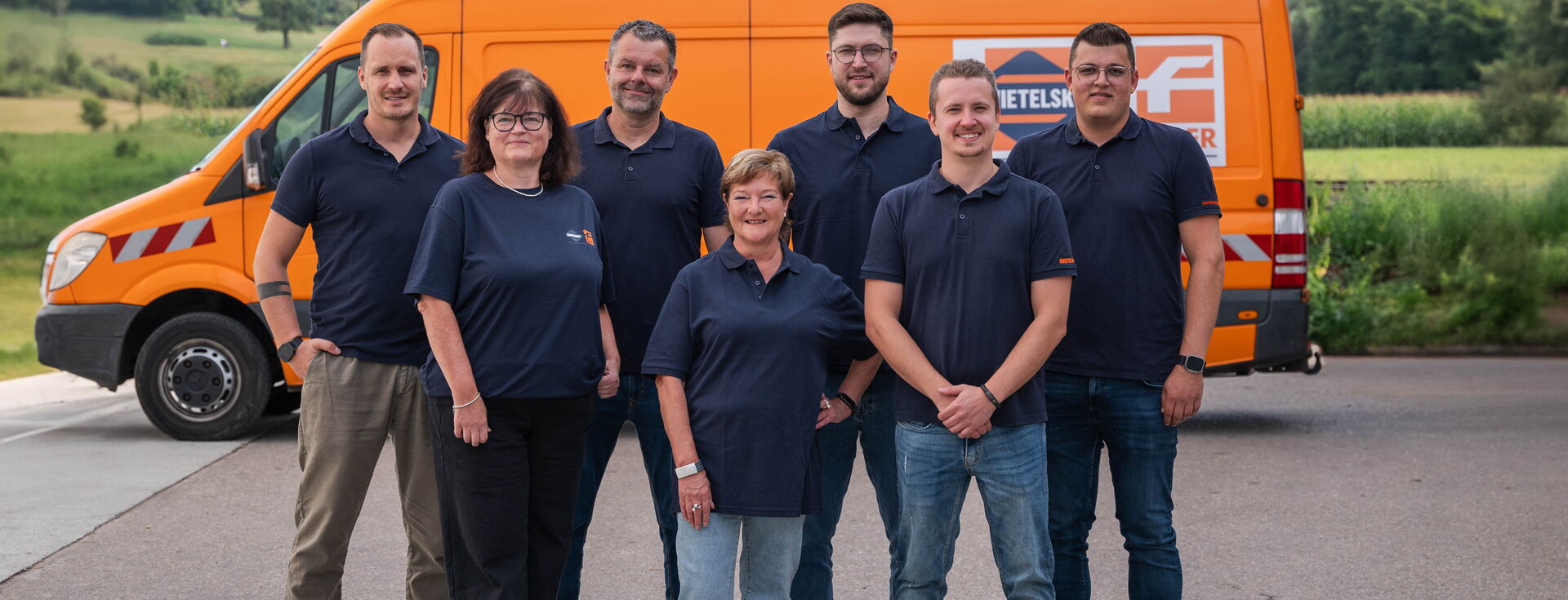 Sieben Personen stehen im Freien vor einem orangefarbenen Servicewagen.