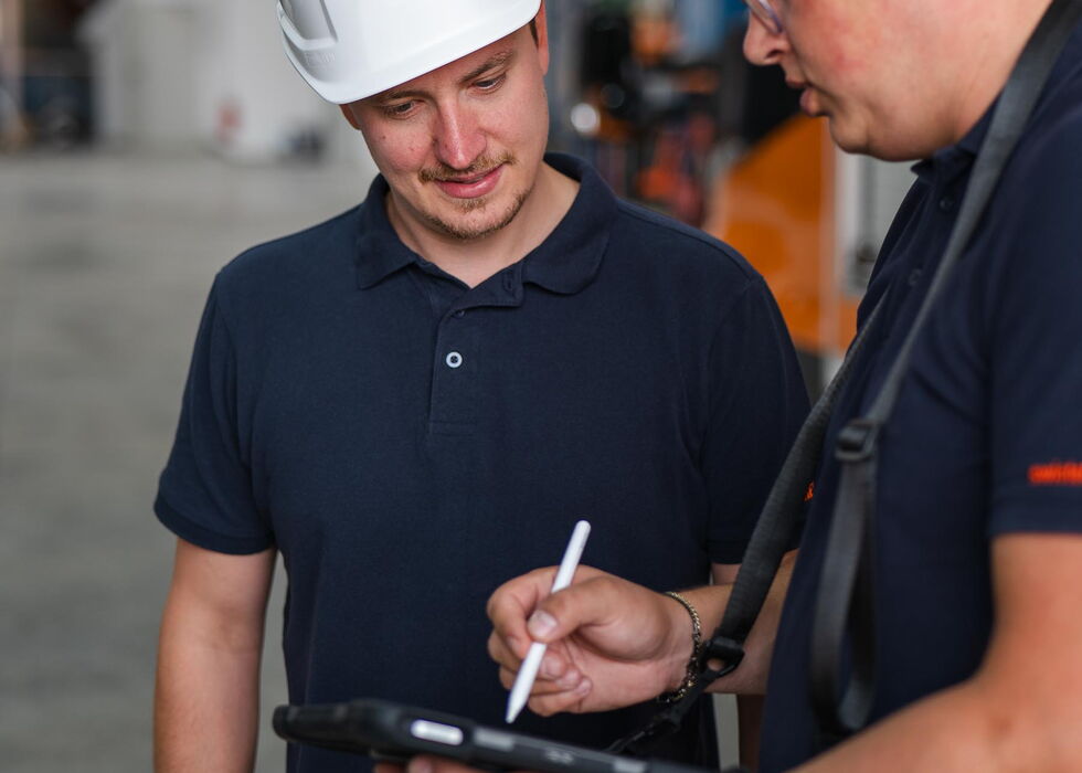 Zwei Lagerarbeiter mit Schutzhelmen prüfen Notizen auf einem Klemmbrett in einem Industriegang.