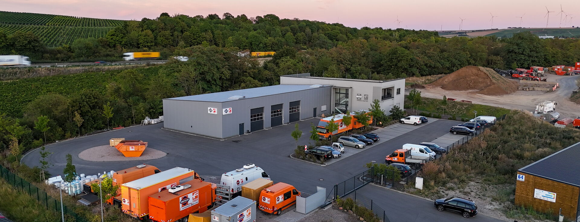 Luftbild eines Industriekomplexes mit weißen Gebäuden, orangen Maschinen und abgestellten Fahrzeugen bei Dämmerung.