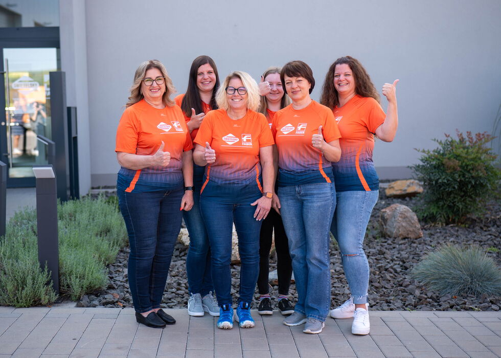 Fünf Frauen stehen lächelnd vor einem Gebäude und tragen orangefarbene T-Shirts und Jeans.