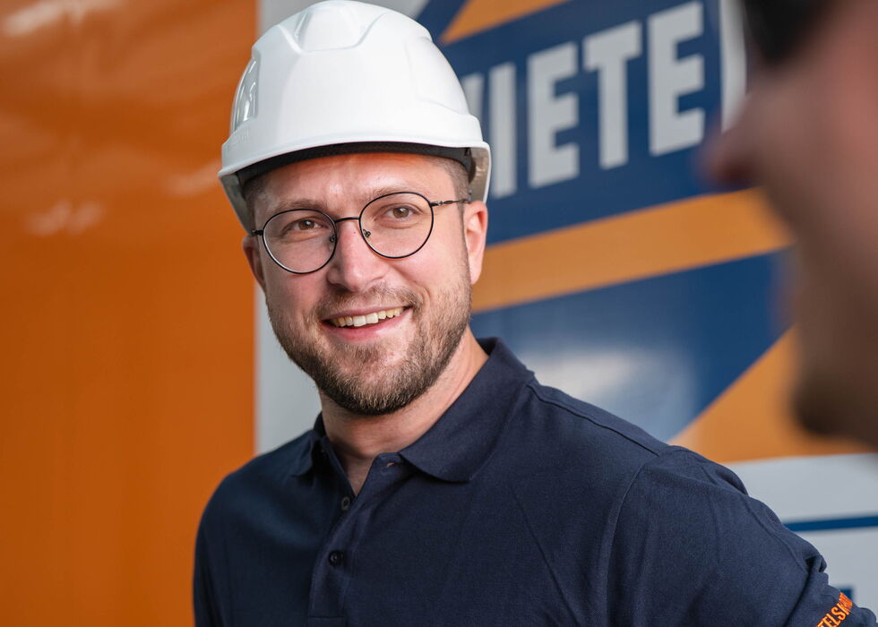 Lächelnder Mann mit weißem Schutzhelm und dunkelblauem Polohemd vor einem orangefarbenen LKW.