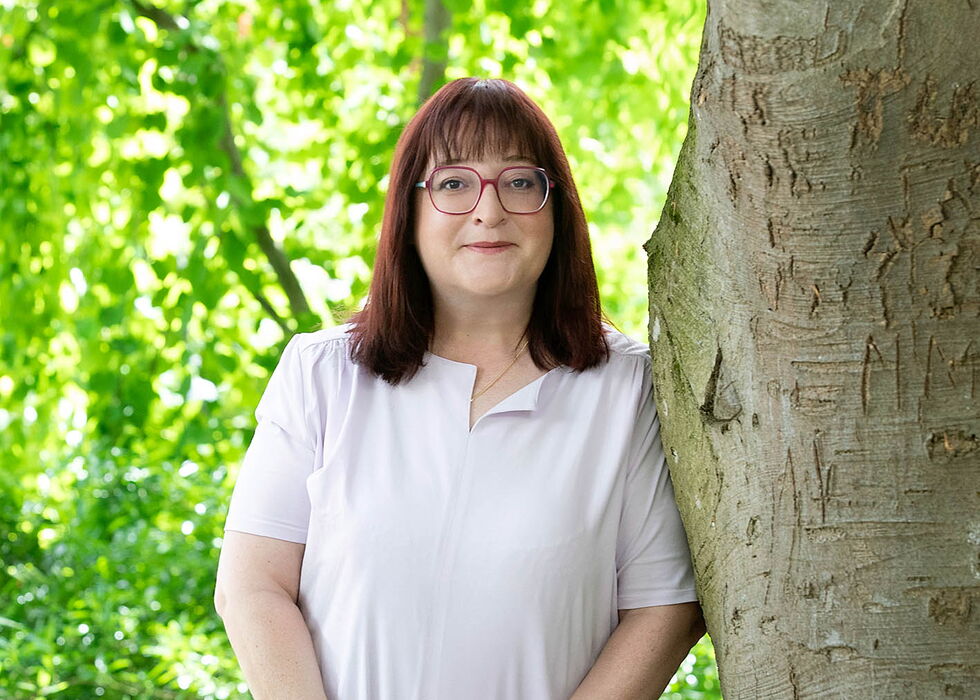 Eine Frau mit Brille steht neben einem Baum in einem grünen Park.