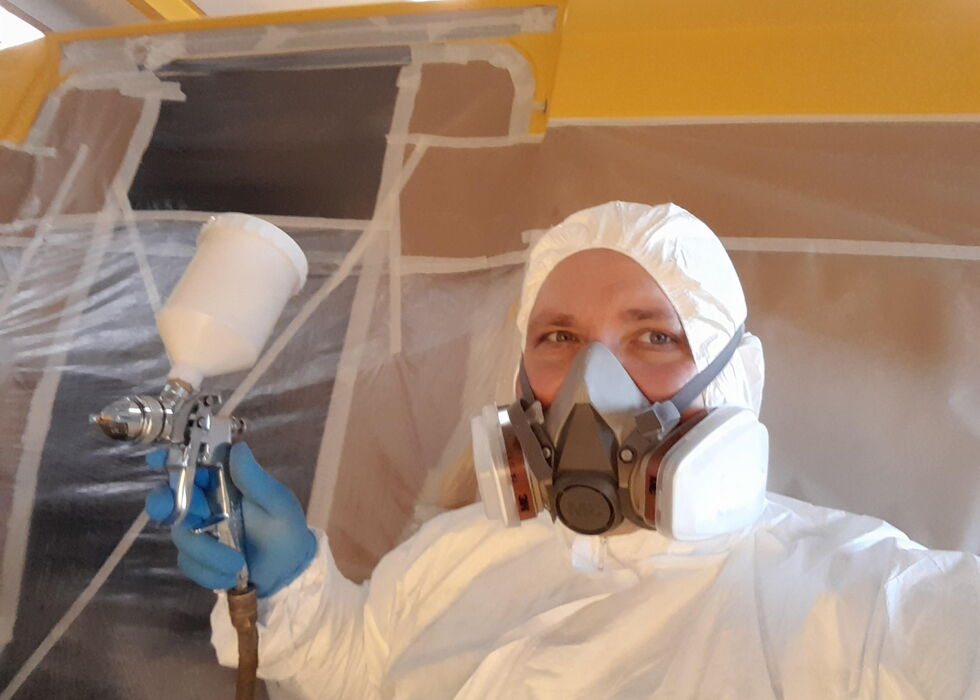 Person in weißem Schutzanzug mit Atemschutzmaske spritzt mit einer Lackierpistole eine Wand im Innenraum.