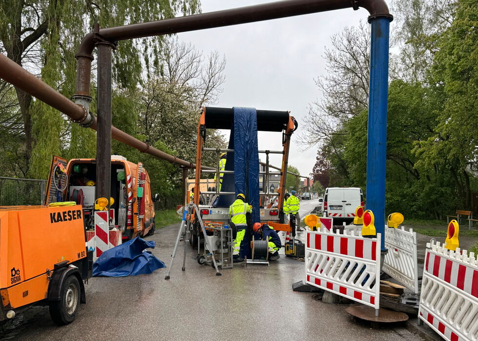 Arbeiter und Absperrungen um Maschinen bei der Reparatur einer Rohrleitungsquerung auf nasser Straße.