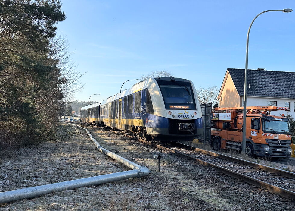 Ein moderner Personenzug fährt auf Bahngleisen vorbei an Häusern und Wartungsfahrzeugen, links daneben ist eine Entwässerungsleitung zu sehen.