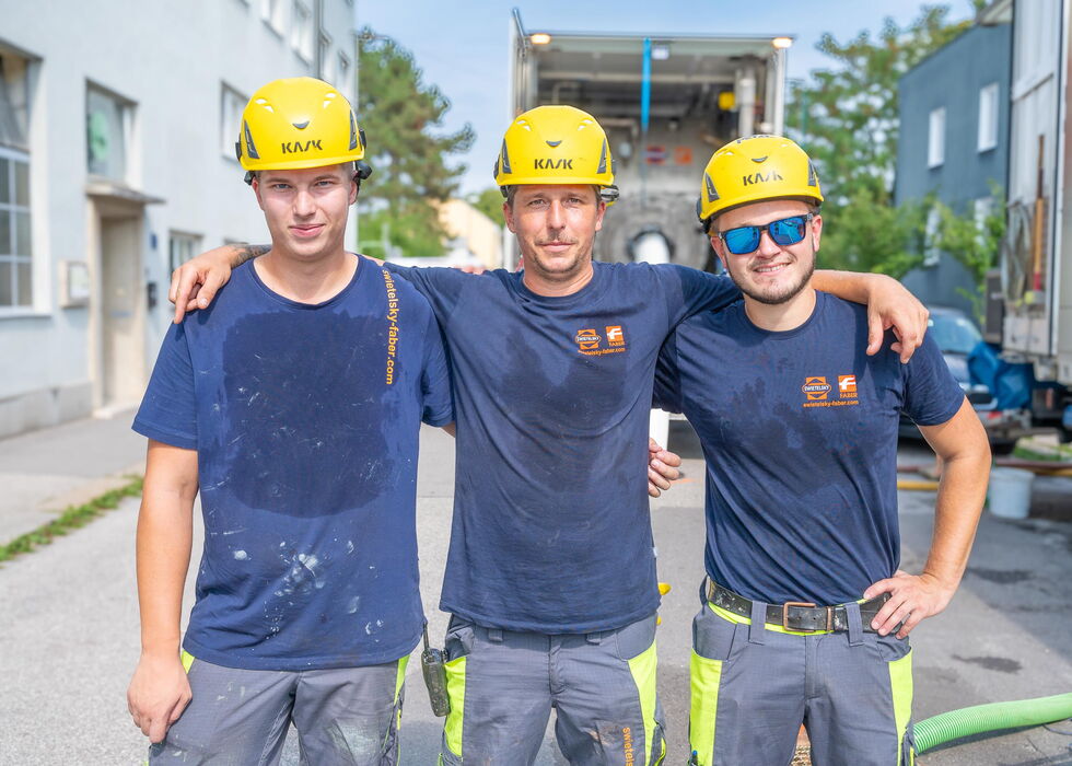 Drei Bauarbeiter mit gelben Helmen stehen gemeinsam im Freien auf einer Baustelle.