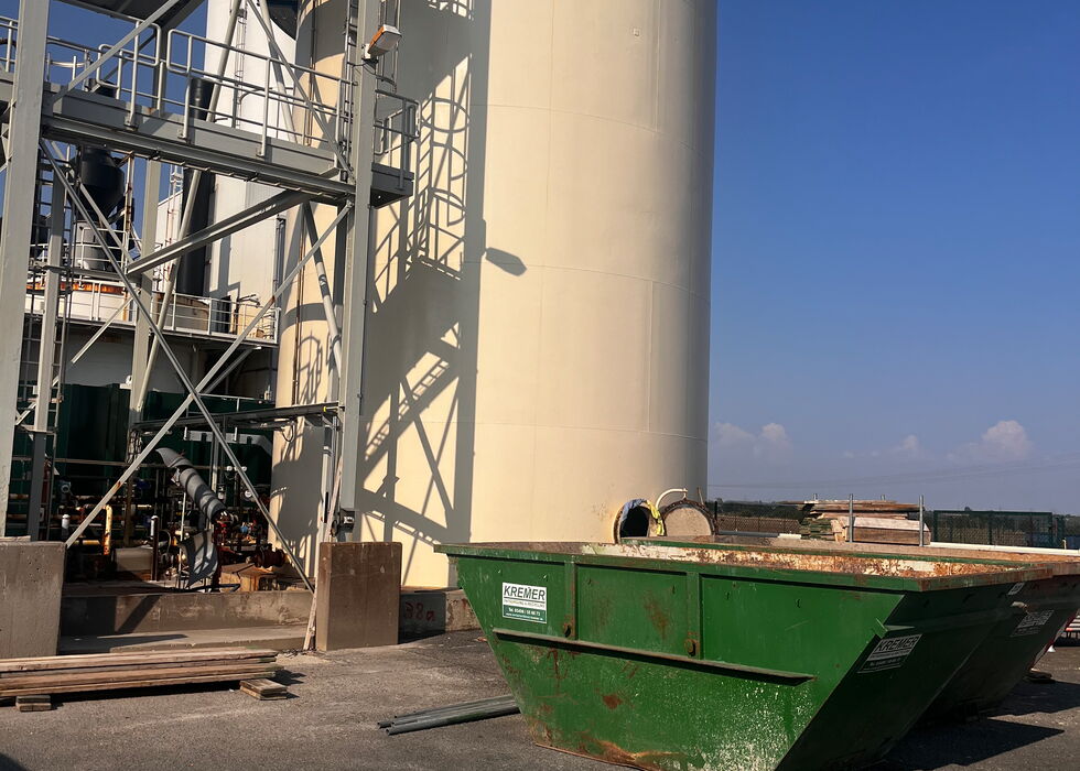 Großer Industriesilo neben Metallkonstruktionen mit einem grünen Abfallcontainer im Vordergrund unter klarem blauem Himmel.
