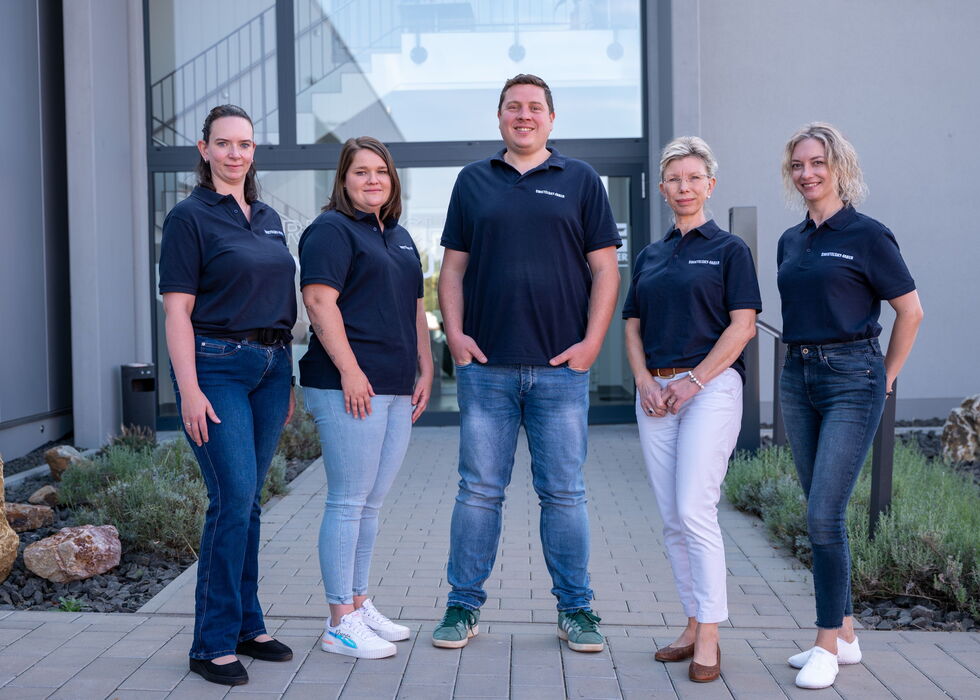 Fünf Teammitglieder im Gesundheitswesen in dunkelblauen Shirts stehen lächelnd auf einem Gehweg im Außenbereich.