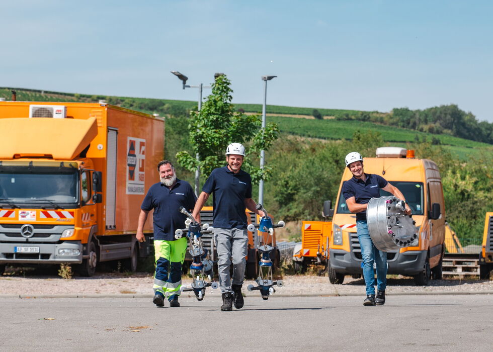 Techniker in Arbeitskleidung stehen mit Roboter-Kameraausrüstung neben orangefarbenen Einsatzfahrzeugen auf einem Außenplatz.