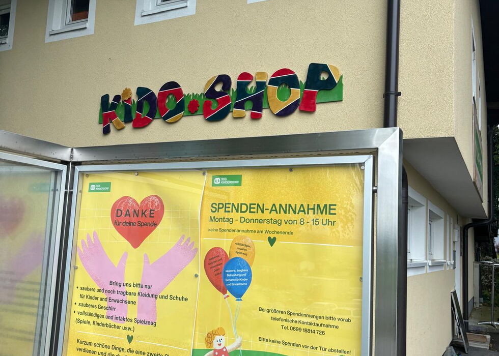 Bunte Spenden-Infotafel vor einem Kinderladen mit gelben Aushängen im Schaufenster.