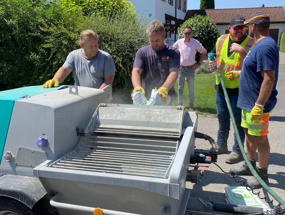 Mehrere Arbeiter mit Handschuhen und Warnwesten reinigen an einer sonnigen Wohnstraße eine mobile Kehrmaschine.