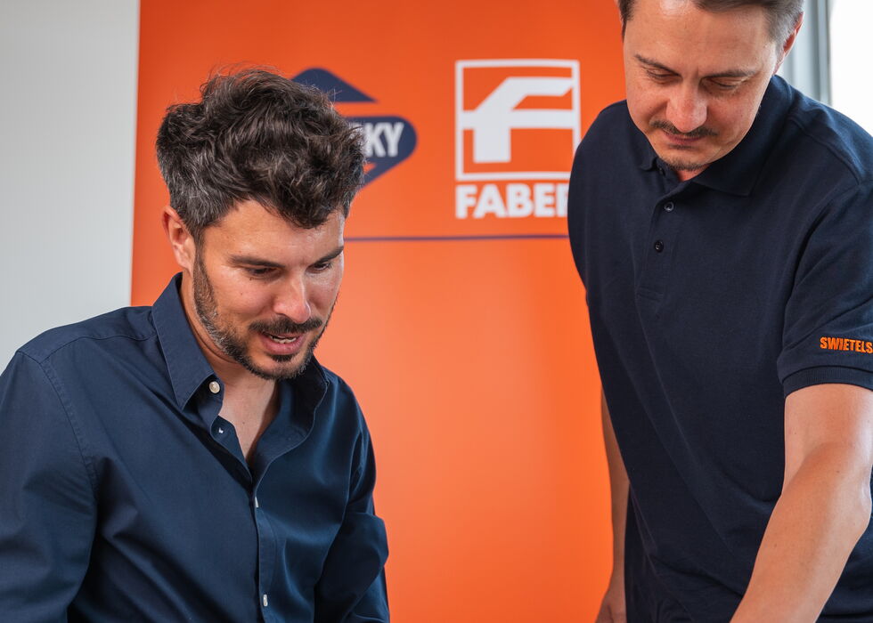 Zwei Männer besprechen im Büro am Schreibtisch Dokumente vor einer orangefarbenen Markenwand.